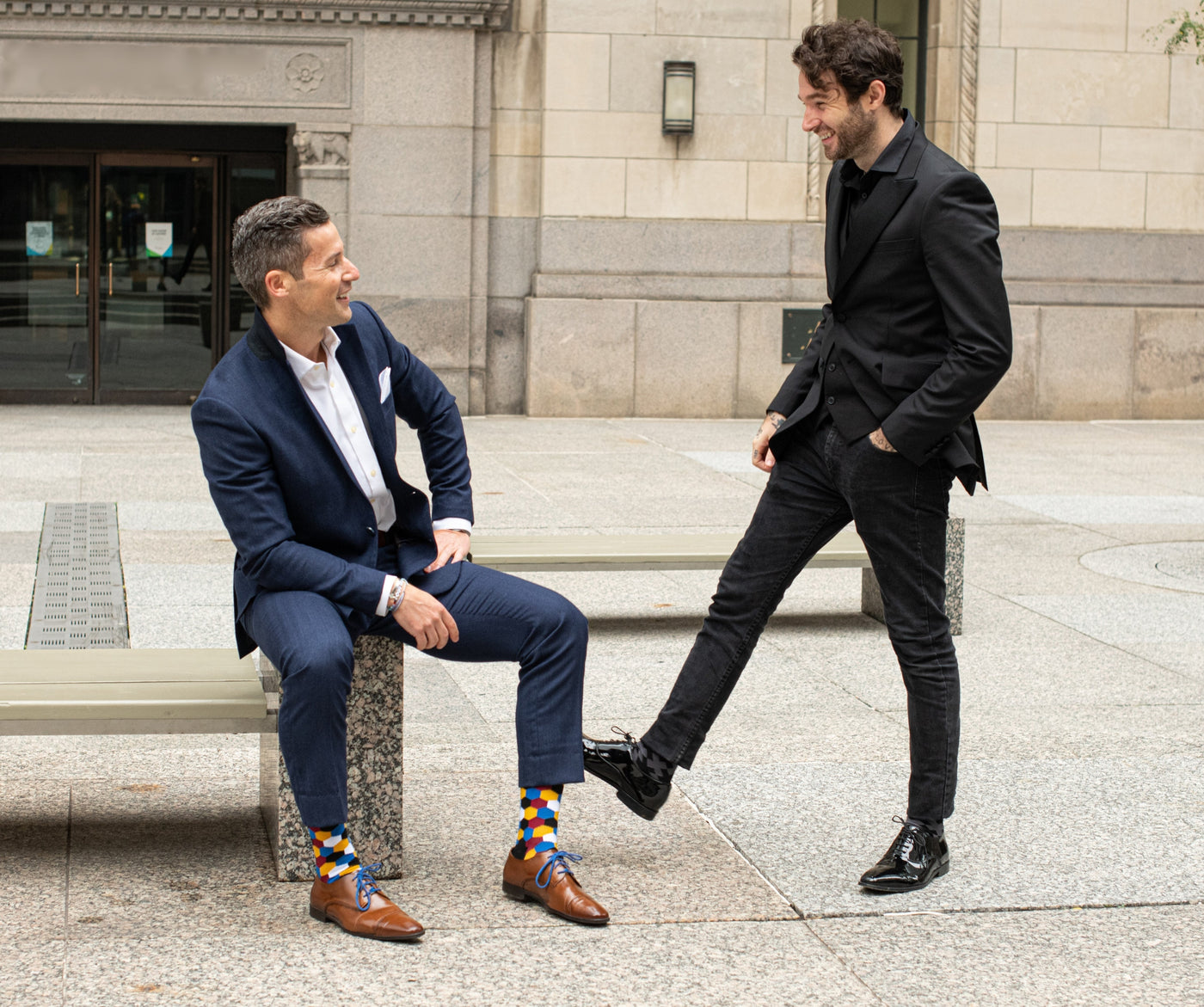two men in suits in front of a building showing off their socks